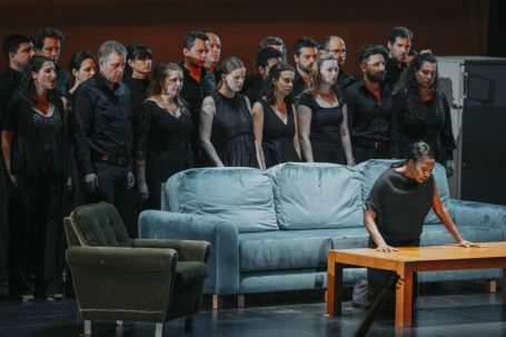Castor et Pollux - Salzburger Festspiele Eine Sängerin sitzt am Tisch, während ein Chor im Hintergrund auftritt.