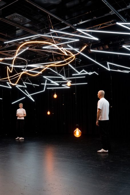 Konstellationen - Rheinisches Landestheater Neuss Zwei Personen in einem dunklen Raum mit leuchtenden Linien und Hängelampen.