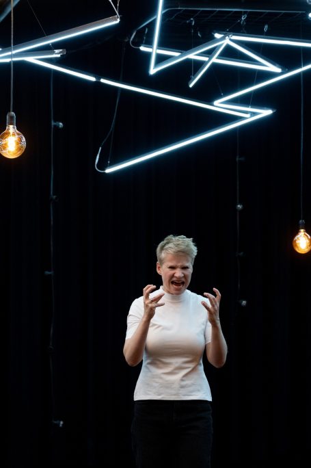 Konstellationen - Rheinisches Landestheater Neuss Eine Person in einem weißen Oberteil steht vor einer dunklen Wand mit geometrischen Lichtern.