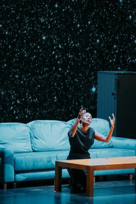 Castor et Pollux - Salzburger Festspiele Eine Tänzerin in schwarzem Kleid, kniend vor einem Tisch, vor einem blauen Sofa.