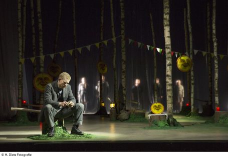 Tannöd - Theater Hof Ein Mann sitzt nachdenklich im Wald, während Figuren im Hintergrund sichtbar sind.
