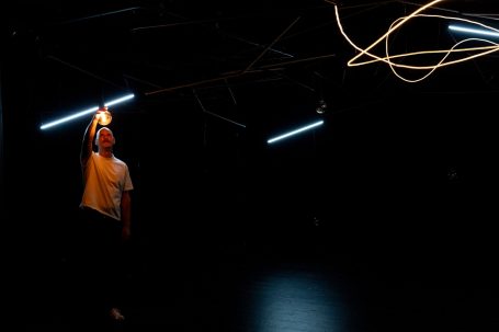 Konstellationen - Rheinisches Landestheater Neuss Eine Person hält ein Licht in der Dunkelheit, umgeben von leuchtenden Linien.